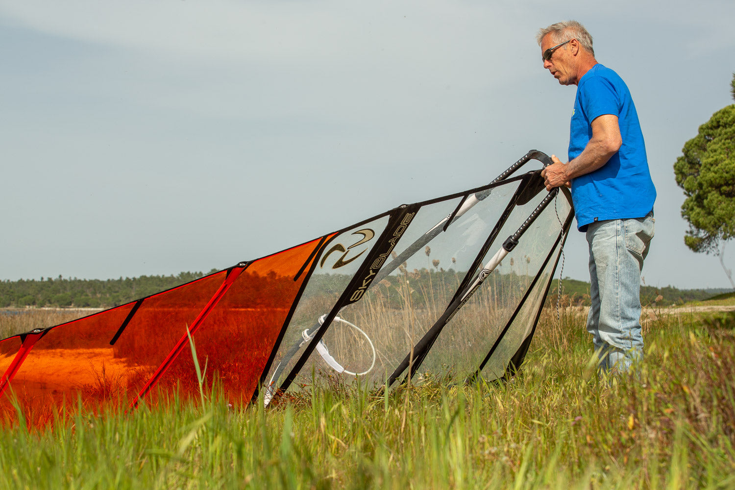 Man setting up a large orange and black windsurf sail in an open field.