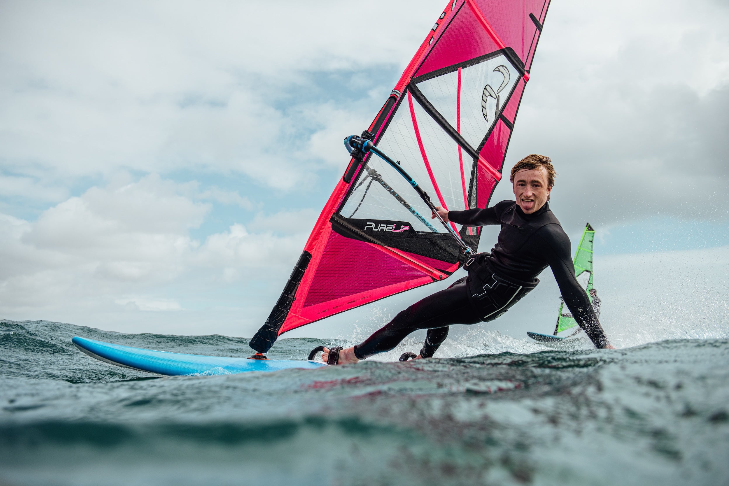 Person windsurfing on a blue board with a pink sail against a cloudy sky.