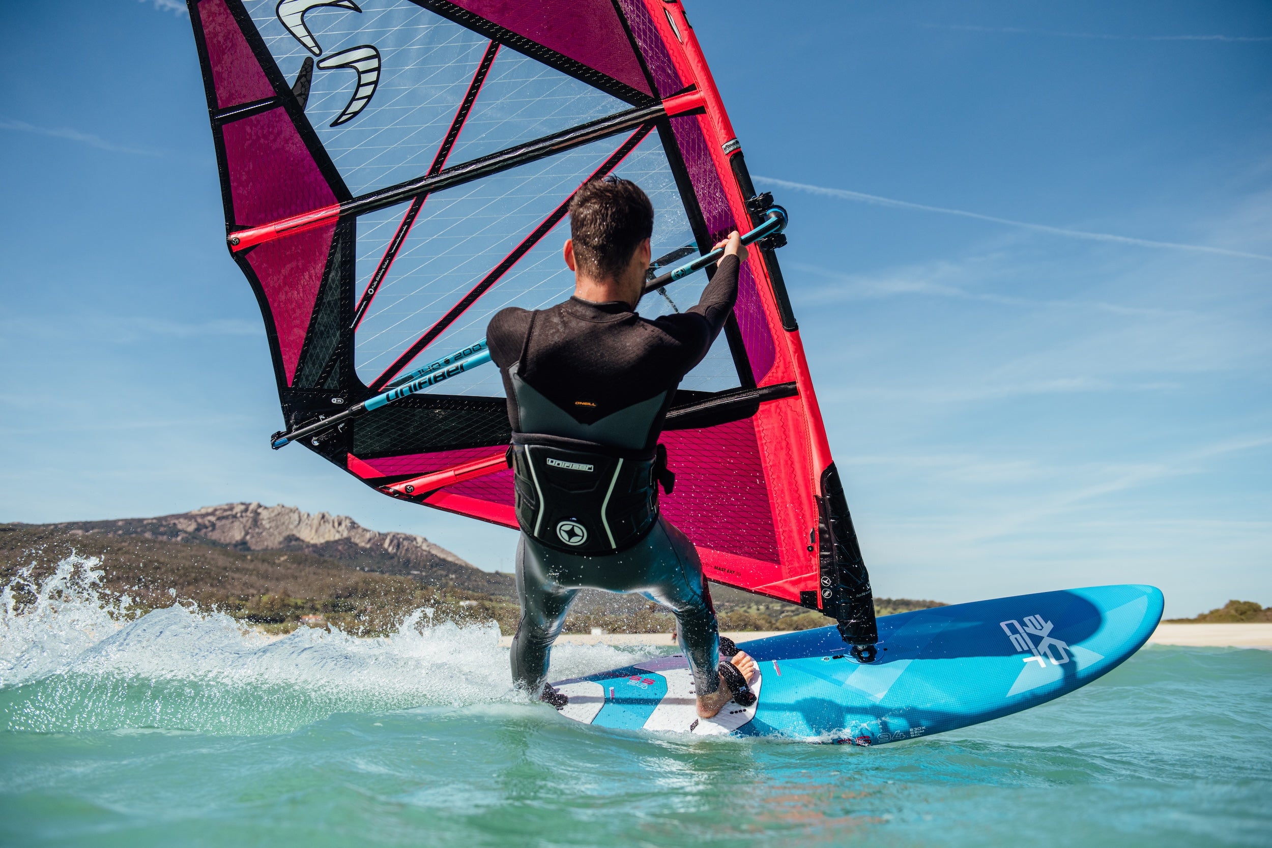 Person windsurfing on a clear day with mountains in the background and a blue board
