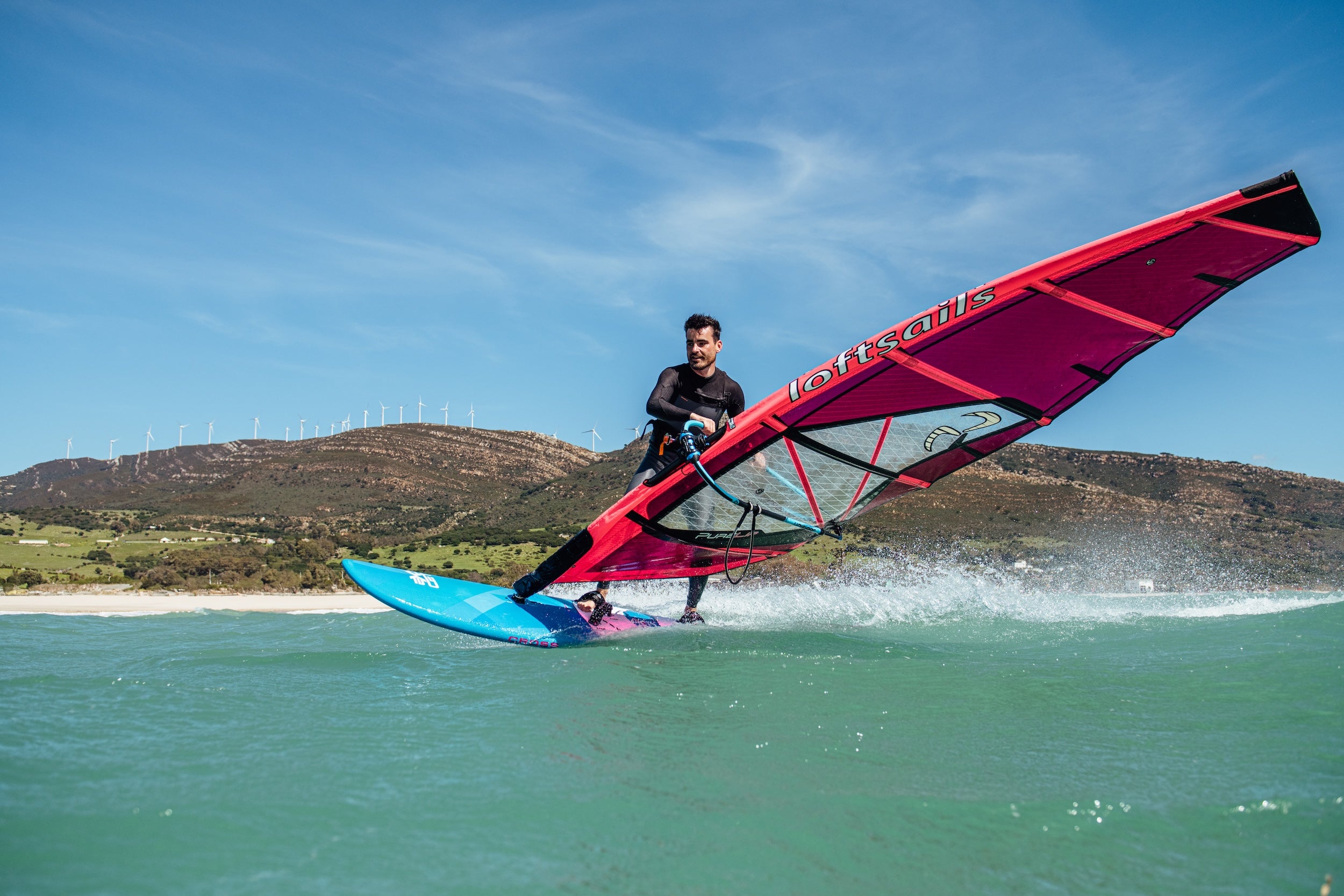 Person windsurfing on a blue and red sail with mountains in the background