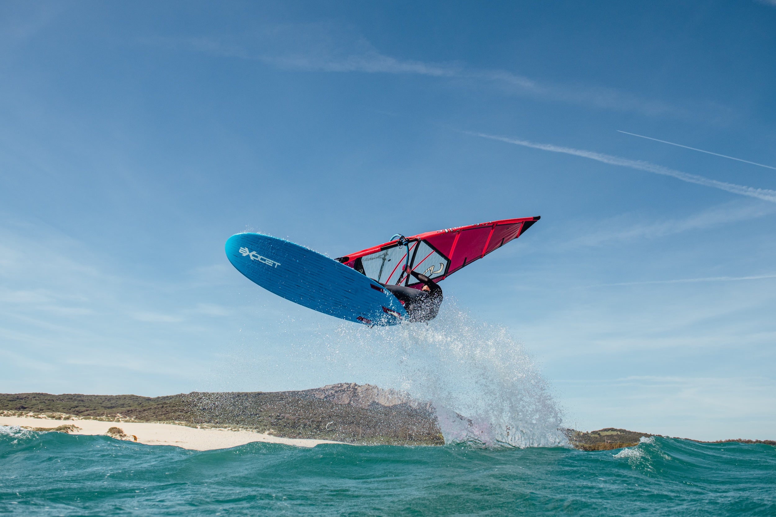 Person windsurfing on a blue board with a red sail against a clear sky.