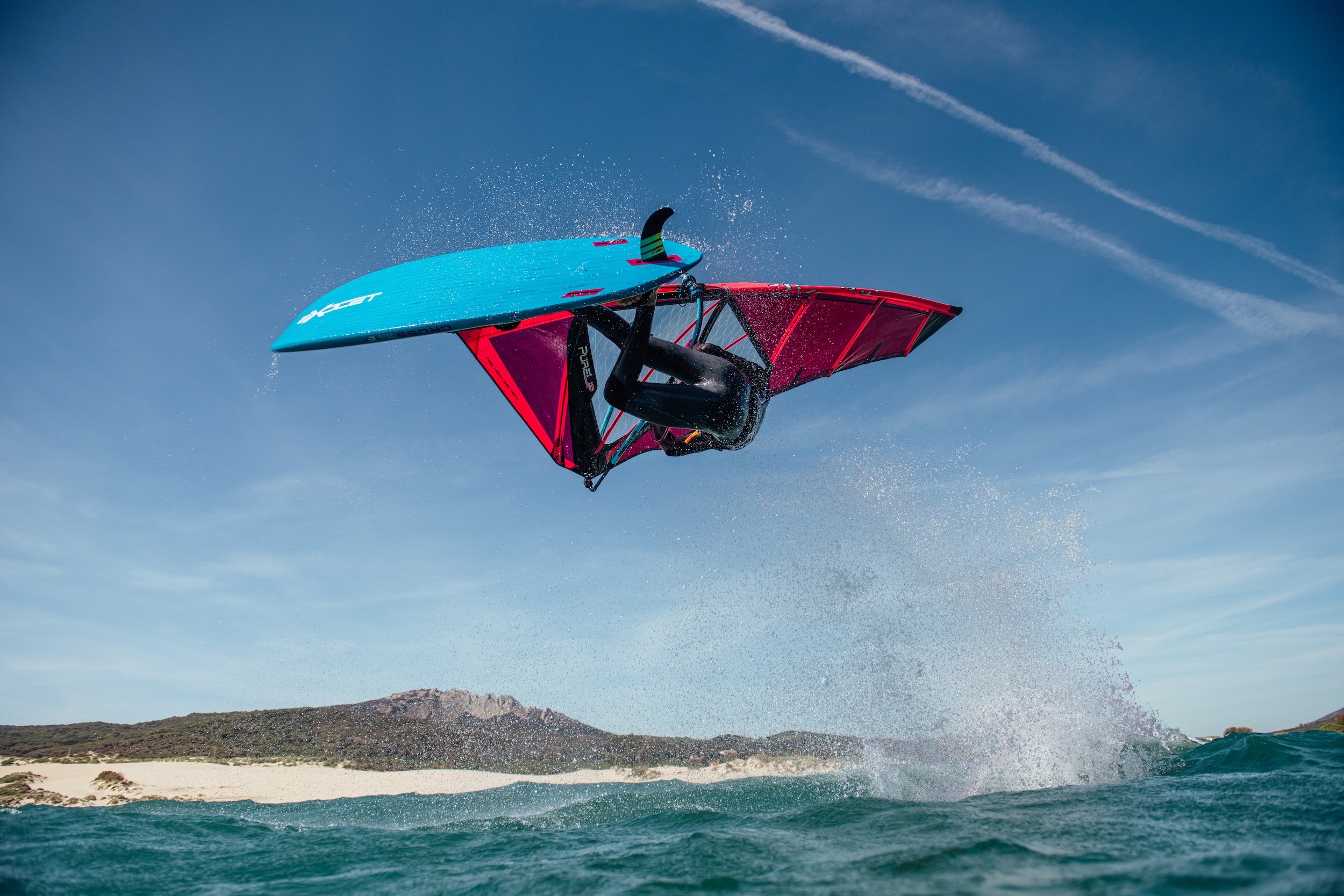 Person on a windsurf board in mid-air over water with a clear blue sky.