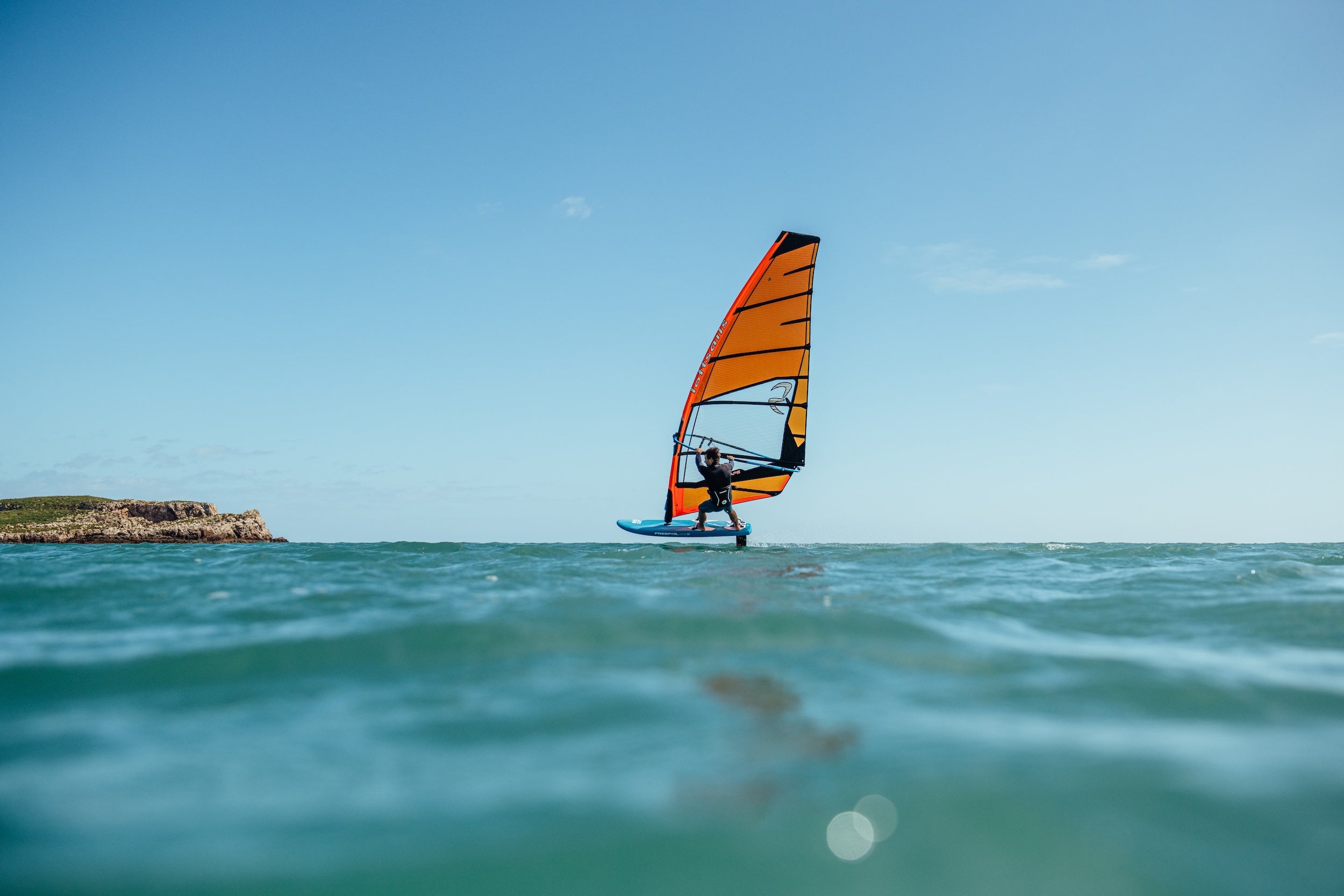 Person windsurfing on a clear day with a bright blue sky and turquoise water.