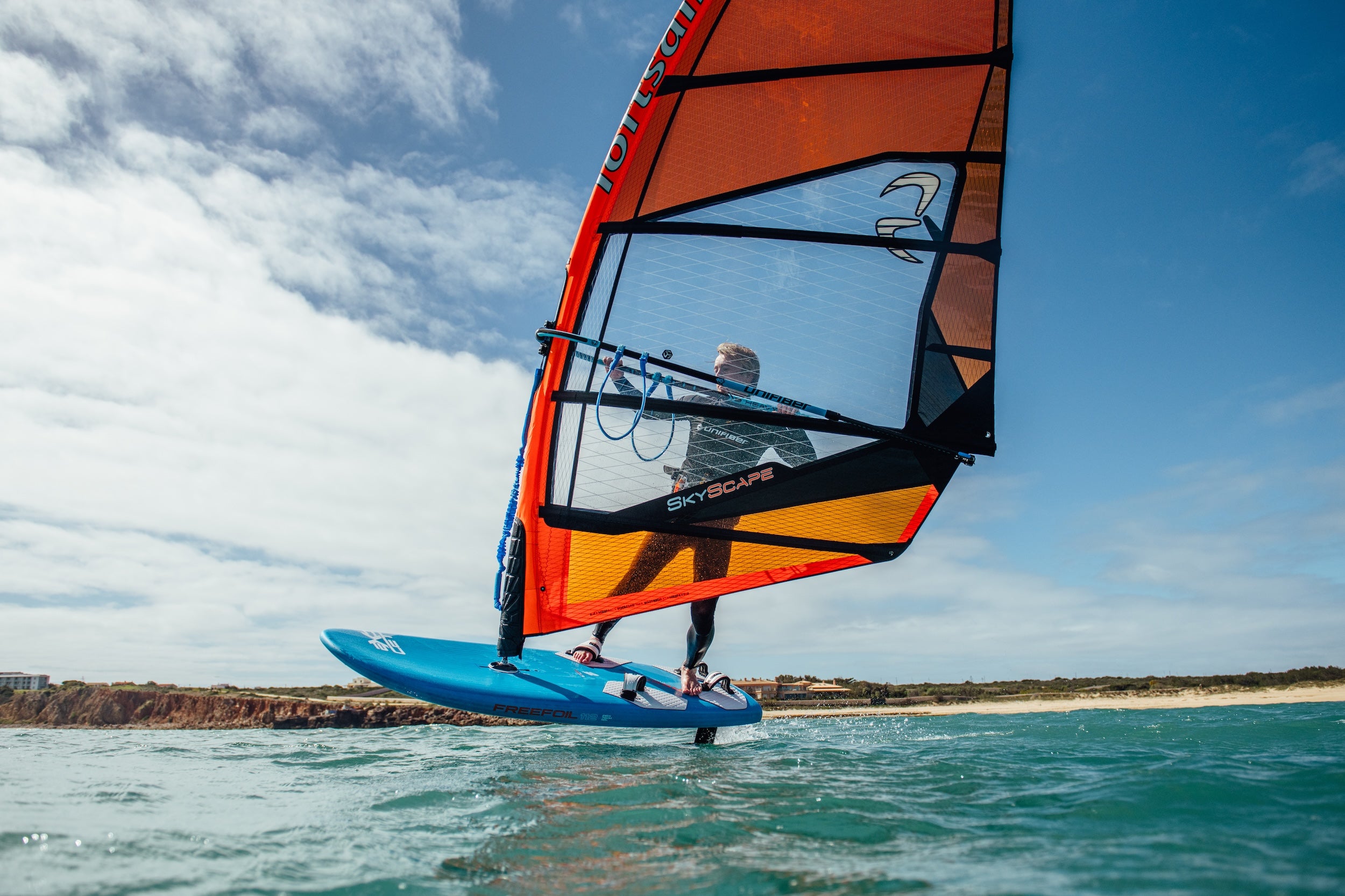 Person windsurfing on a blue board with an orange sail against a clear sky.