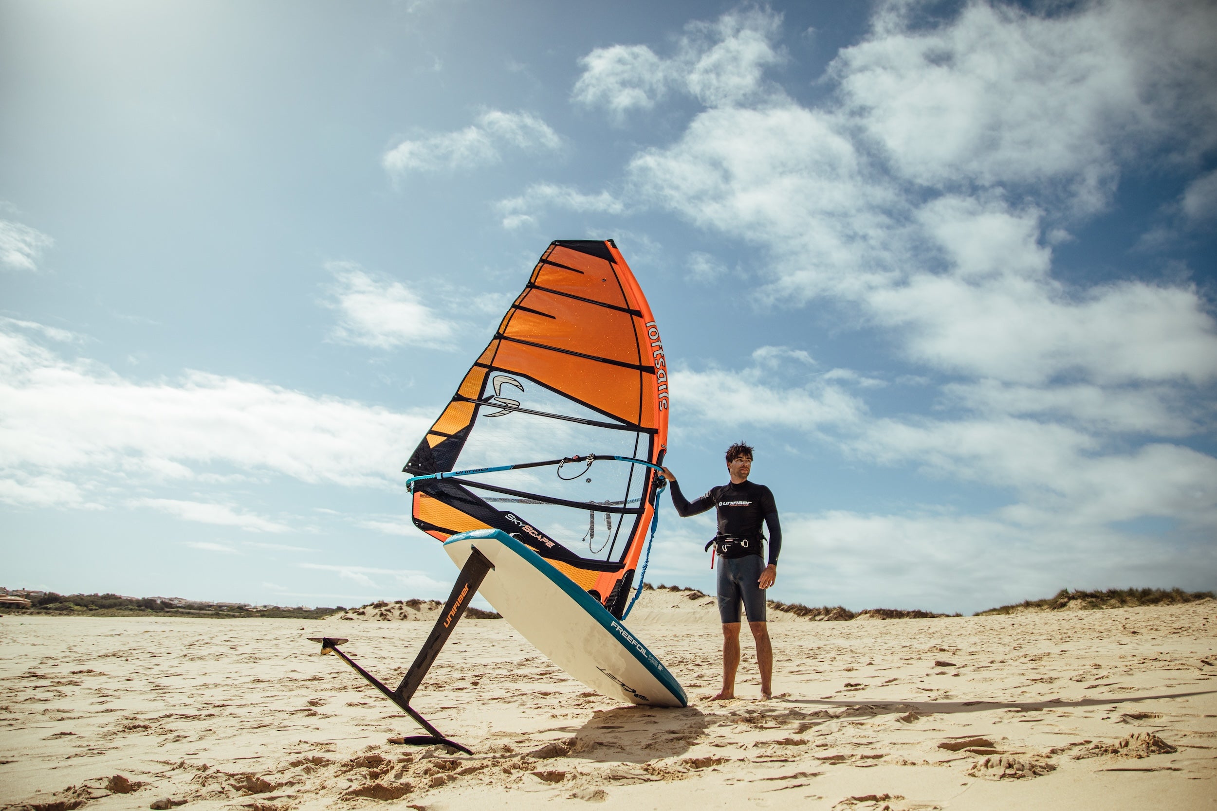 Person holding a windsurf board on a sandy beach with a blue sky.