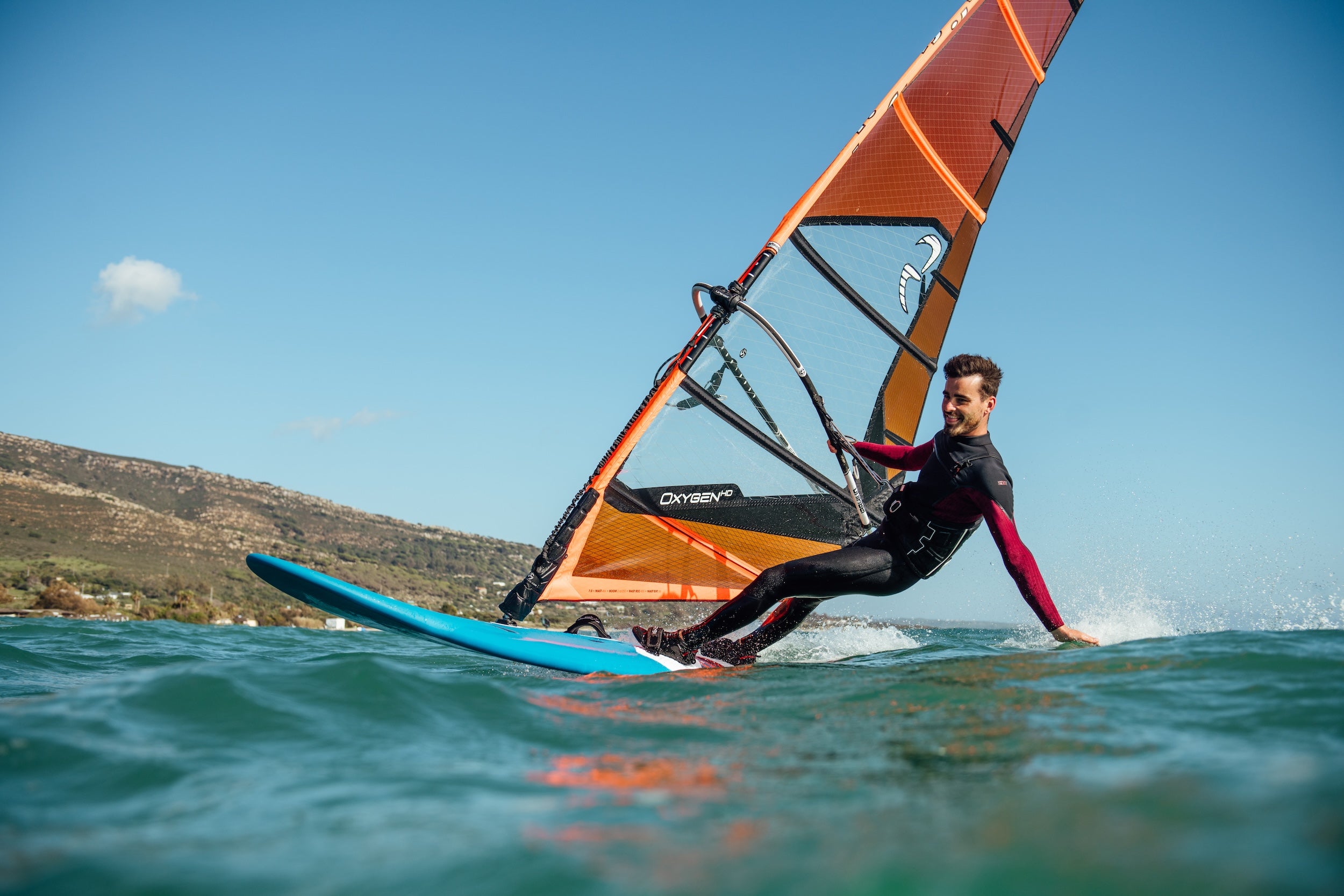 Person windsurfing on a blue board with an orange sail against a clear sky.