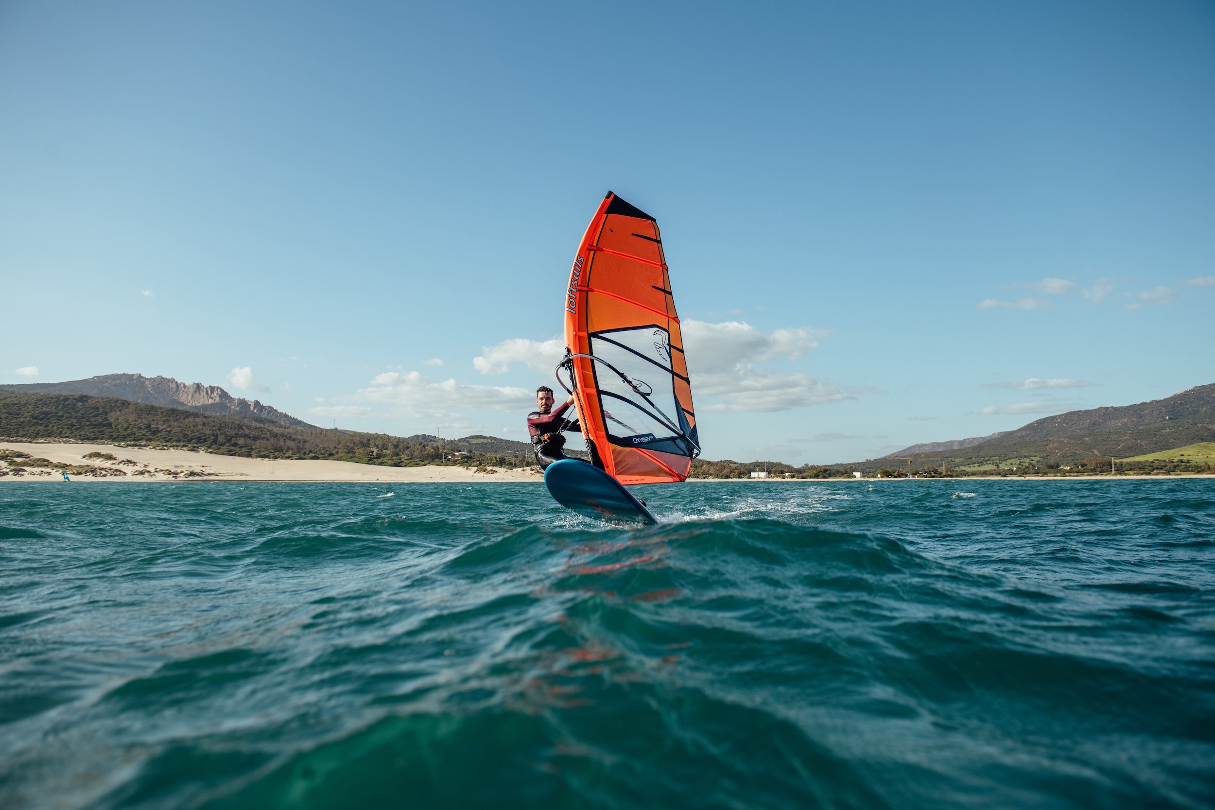 Person windsurfing on a blue and orange sail with a scenic background of mountains and beach.