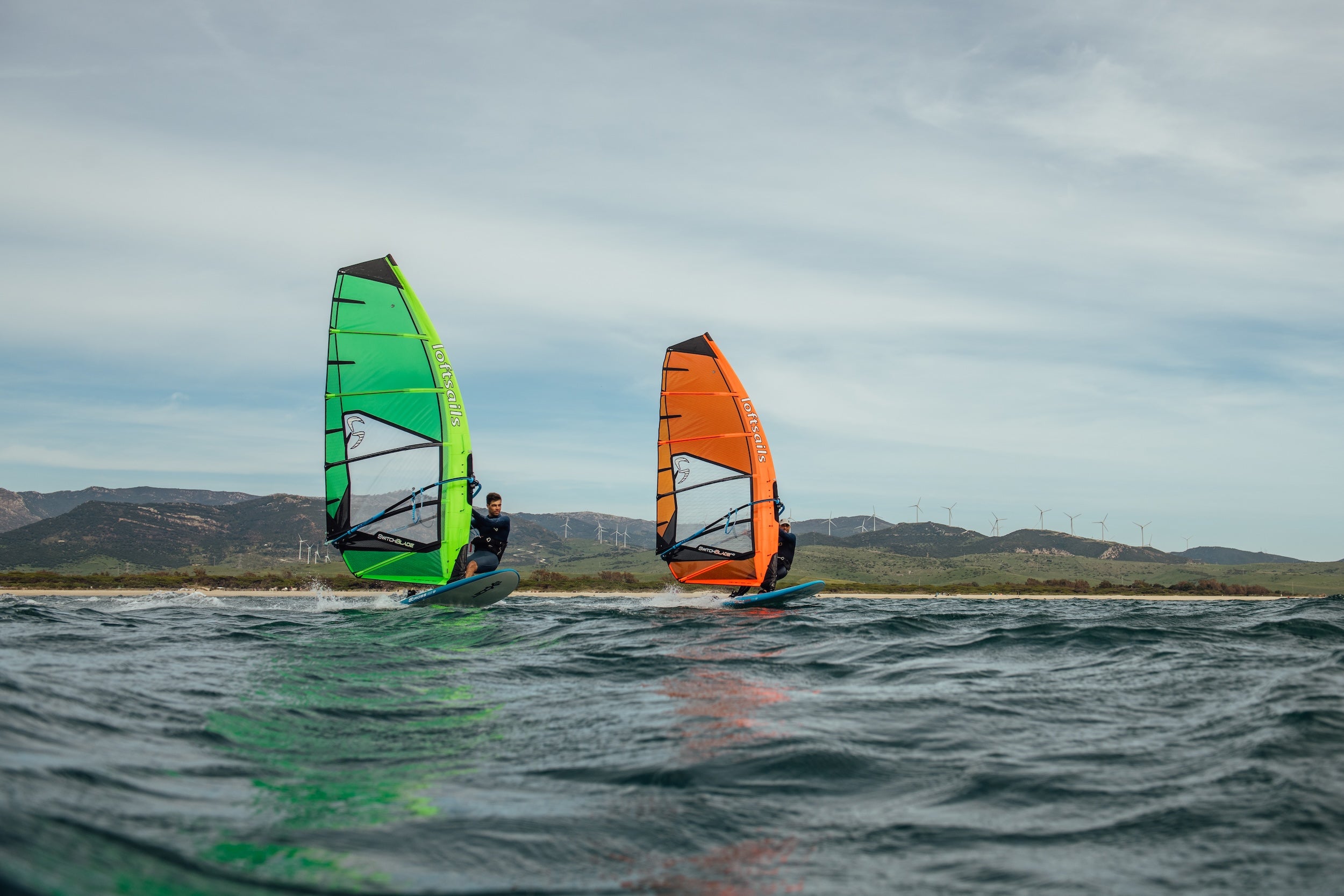 Two windsurfers on the water with colorful sails against a cloudy sky.