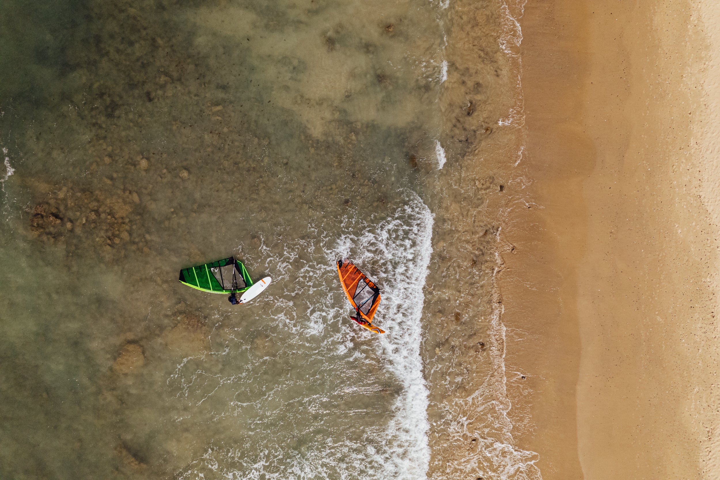 Two windsurf board on a beach with waves lapping at their sides, viewed from above.