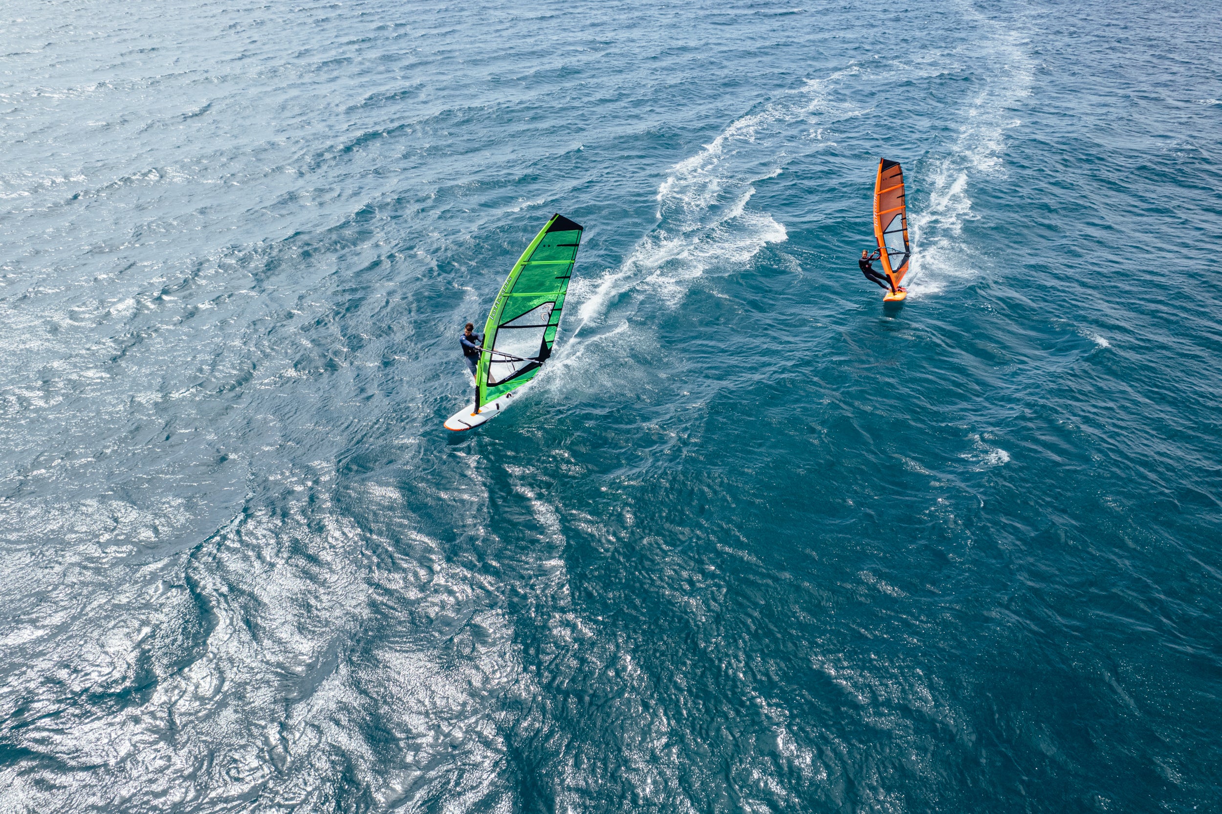 Two windsurfers on the water with one using a green sail and the other an orange sail.