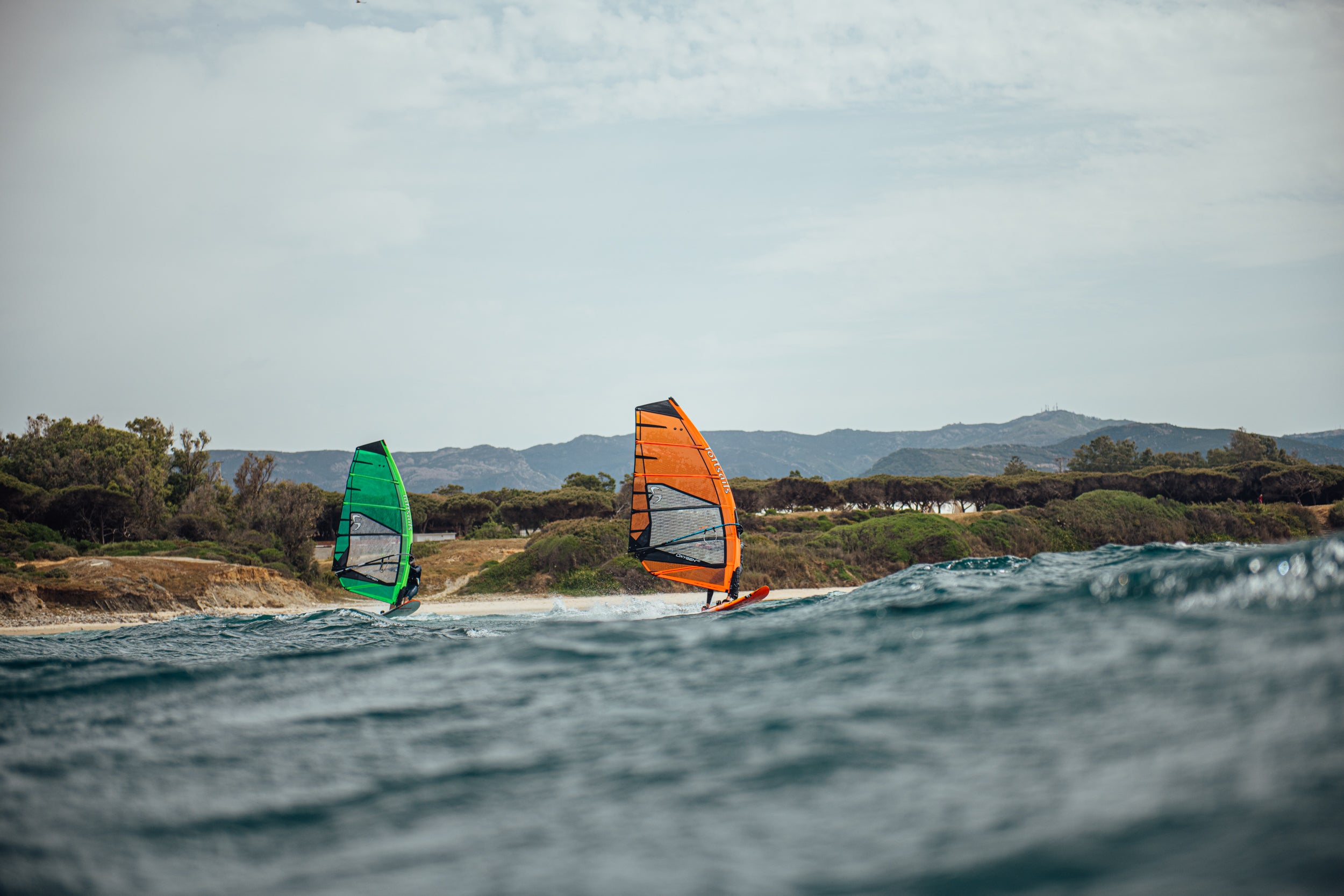 Two windsurfers with colorful sails on a body of water with a scenic background.