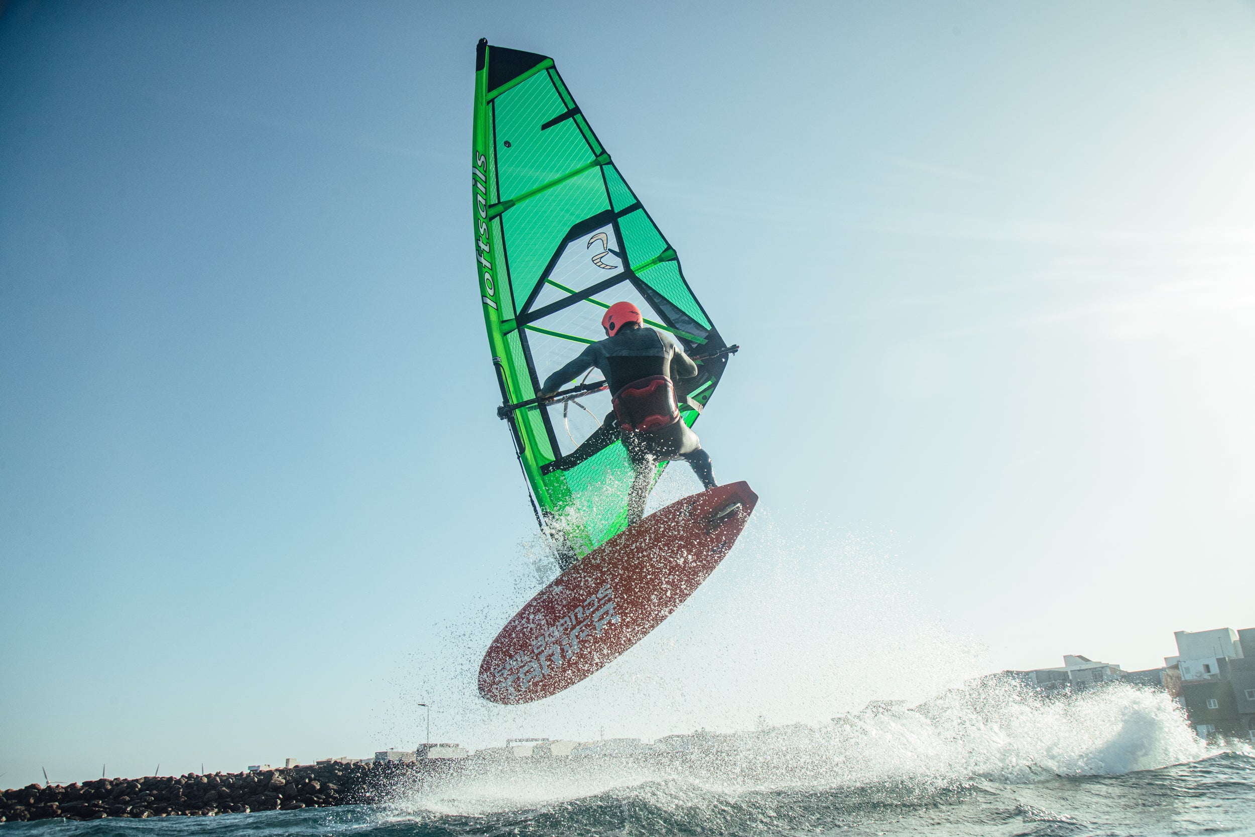 Person windsurfing with a green sail and red helmet against a clear blue sky.
