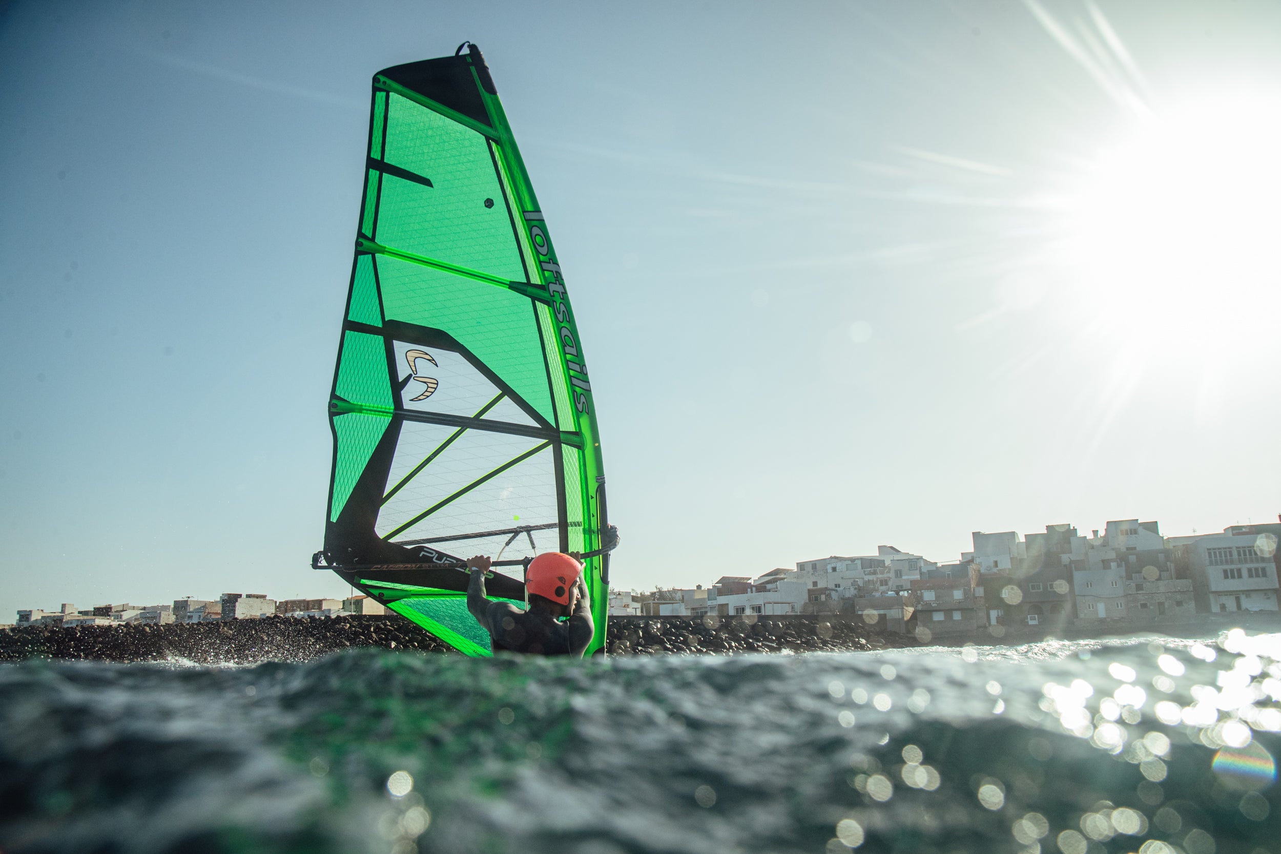 Windsurfing sail with a green and black design on the water, with buildings in the background.