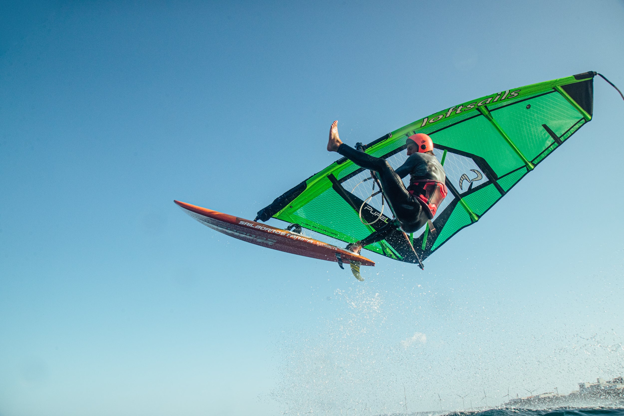 Person windsurfing with a green sail and orange board against a clear blue sky.