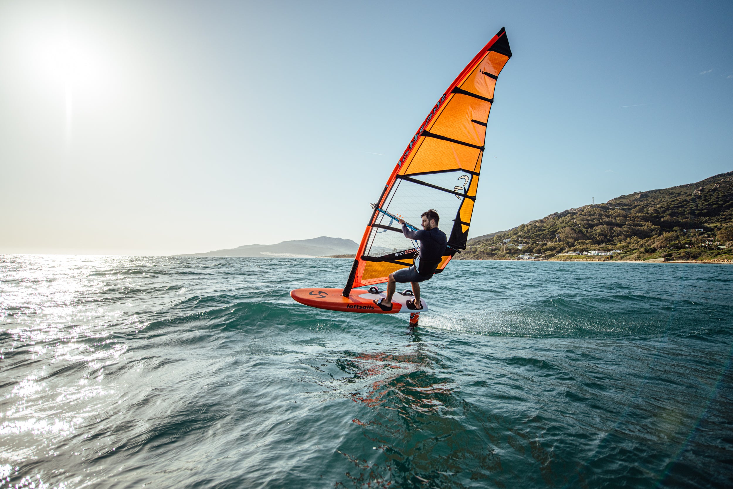 Person windsurfing on a sunny day with a clear blue sky and mountains in the background.