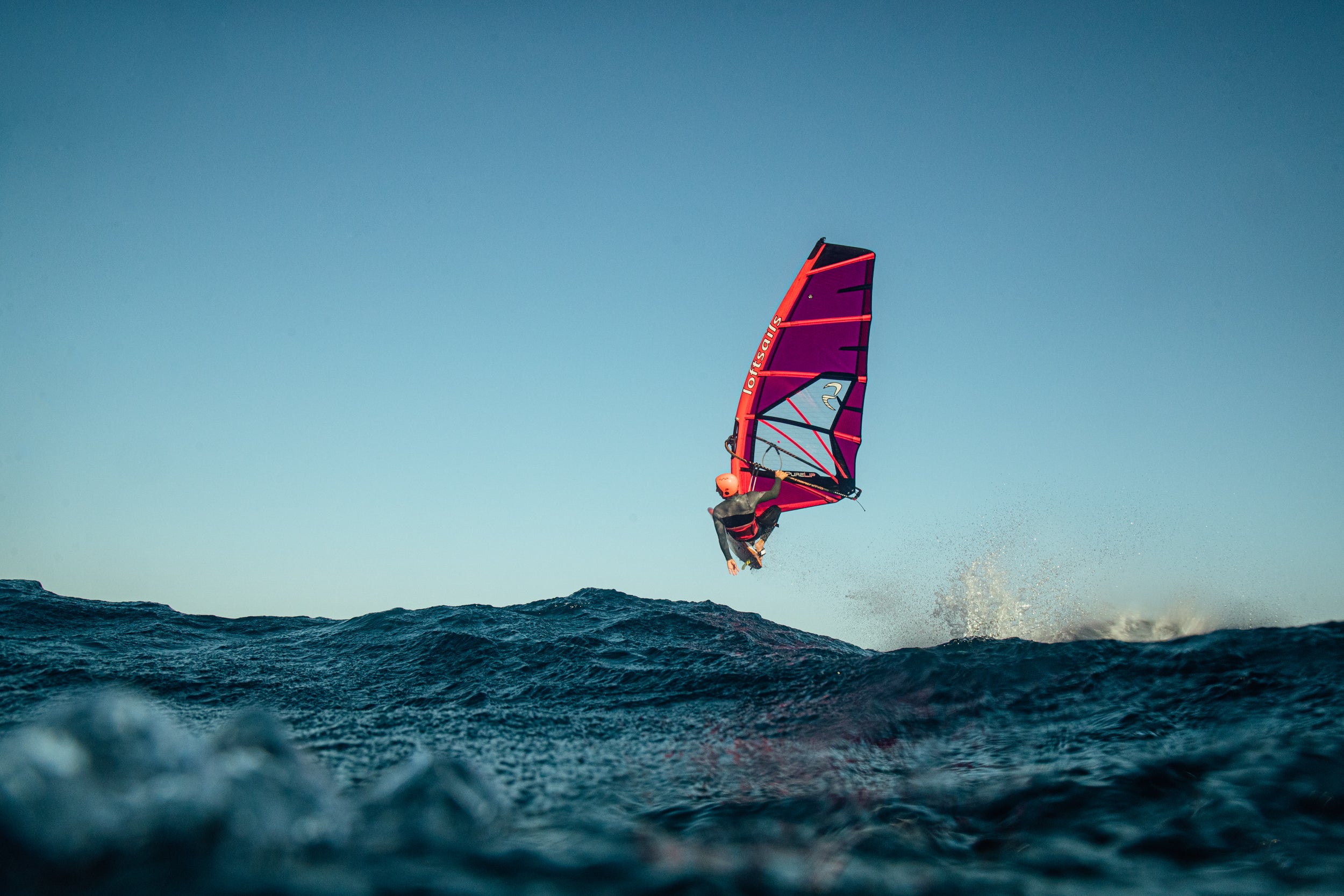 Windsurfer with a red sail gliding over the ocean against a clear blue sky.